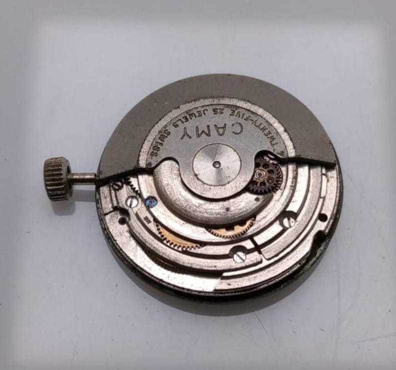 May include: A close-up of a watch movement with the brand name "CAMY" visible. The movement is silver-toned with intricate gears, screws, and a winding stem. The text "TWENTY-FIVE JEWELS SWISS" is also present.