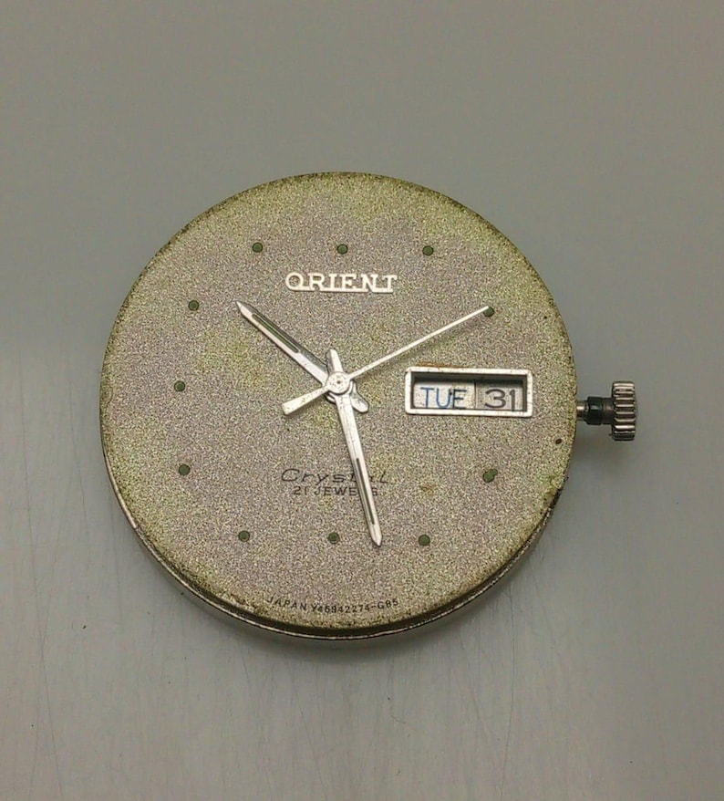 May include: A vintage Orient watch face with a textured, gold-toned dial. The watch face displays the Orient logo, the day and date (TUE 31), and silver hands. The watch has a crown and is marked "Crystal 21 Jewels" and "Japan Y46942274-G05".