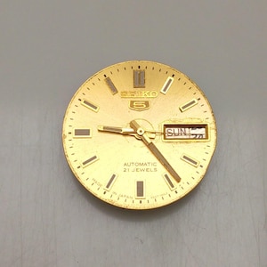 May include: A gold-toned Seiko 5 watch face with a textured finish. The watch face features black rectangular hour markers, gold hands, and a day-date display. The text "SEIKO 5", "AUTOMATIC 21 JEWELS", and "MADE IN JAPAN" are visible.