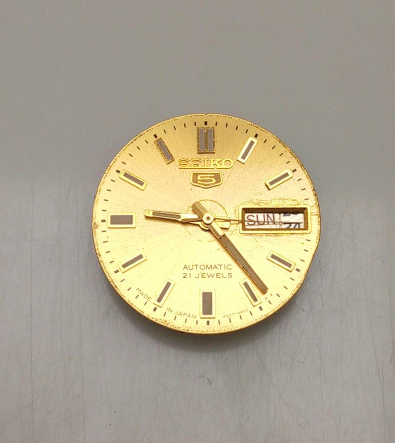 May include: A gold-toned Seiko 5 watch face with a textured finish. The watch face features black rectangular hour markers, gold hands, and a day-date display. The text "SEIKO 5", "AUTOMATIC 21 JEWELS", and "MADE IN JAPAN" are visible.
