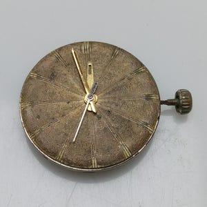 Op de afbeelding: Een goudkleurig horloge-wijzerplaat met een cirkelvormig, getextureerd ontwerp en goudkleurige wijzers. De wijzerplaat is bevestigd aan een kleine, goudkleurige steel.