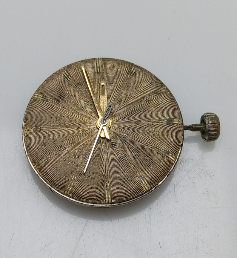 Op de afbeelding: Een goudkleurig horloge-wijzerplaat met een cirkelvormig, getextureerd ontwerp en goudkleurige wijzers. De wijzerplaat is bevestigd aan een kleine, goudkleurige steel.