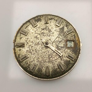 May include: A vintage watch face with a distressed, metallic finish. The face features the brand name "Gandino" and the text "Swiss Made". The watch face has rectangular hour markers and a date window displaying the number "4".