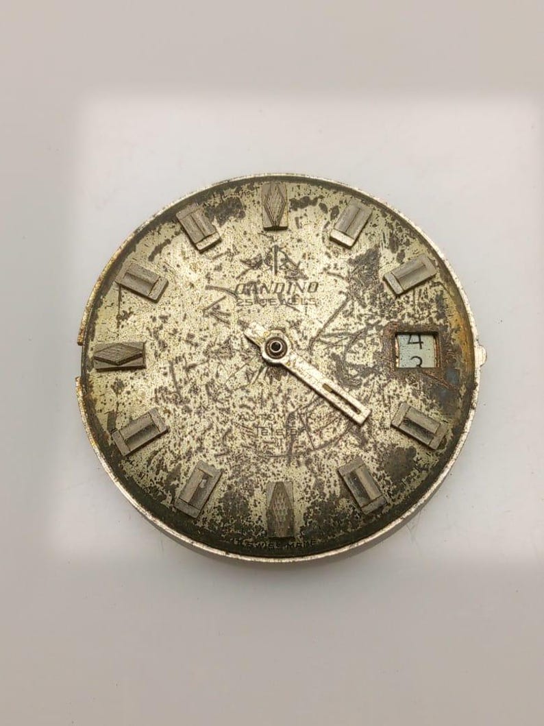 May include: A vintage watch face with a distressed, metallic finish. The face features the brand name "Gandino" and the text "Swiss Made". The watch face has rectangular hour markers and a date window displaying the number "4".