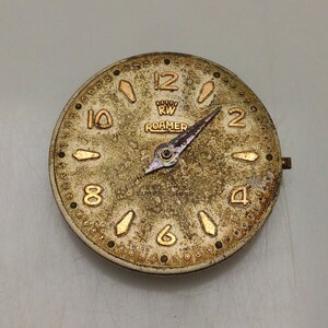 May include: Vintage Roamer watch face with a gold-toned, textured surface. The face features gold numerals, markers, and hands. The text "RW ROAMER" is visible above the center. The watch face has a small, protruding stem on the right side.