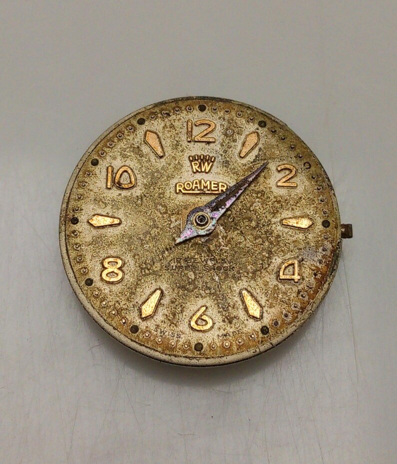 May include: Vintage Roamer watch face with a gold-toned, textured surface. The face features gold numerals, markers, and hands. The text "RW ROAMER" is visible above the center. The watch face has a small, protruding stem on the right side.