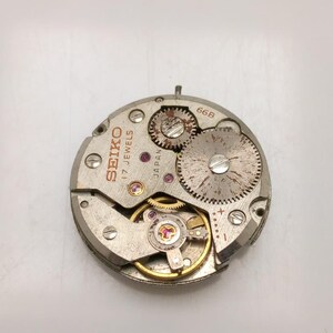 May include: A close-up of a Seiko watch movement with visible gears, jewels, and the text "SEIKO 17 JEWELS JAPAN". The silver-toned mechanism has a circular shape with various components and screws. The watch movement is a vintage item.