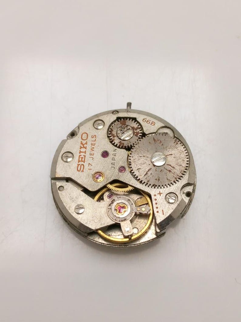 May include: A close-up of a Seiko watch movement with visible gears, jewels, and the text "SEIKO 17 JEWELS JAPAN". The silver-toned mechanism has a circular shape with various components and screws. The watch movement is a vintage item.