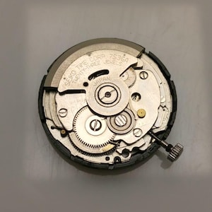 May include: A close-up of a Seiko Time Corp 7S36A watch movement. The silver-colored mechanism features numerous gears, jewels, and a winding stem. The text "JAPAN" is visible on the movement.