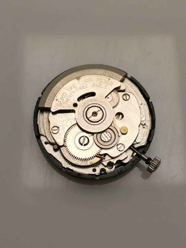 May include: A close-up of a Seiko Time Corp 7S36A watch movement. The silver-colored mechanism features numerous gears, jewels, and a winding stem. The text "JAPAN" is visible on the movement.