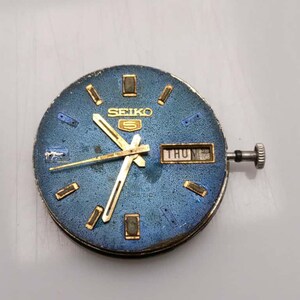 May include: A vintage Seiko watch face with a blue dial and gold-tone hands and markers. The watch face displays the day of the week and the Seiko logo. The watch has a silver crown and is an automatic watch.