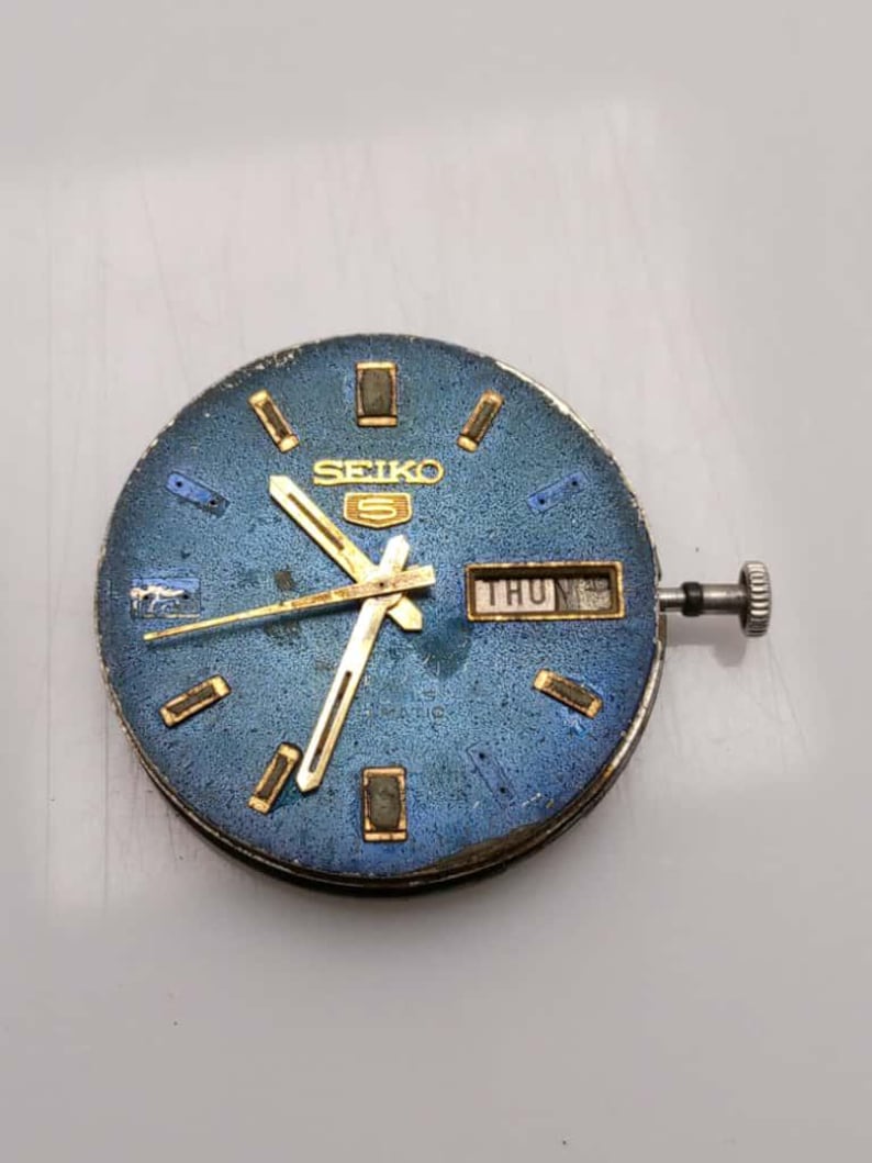 May include: A vintage Seiko watch face with a blue dial and gold-tone hands and markers. The watch face displays the day of the week and the Seiko logo. The watch has a silver crown and is an automatic watch.