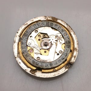 May include: A detailed view of a watch movement, showcasing intricate gears, springs, and a date dial. The circular mechanism features a gold-toned outer ring and a silver inner dial with numbered markings. The watch components are set against a grey background.