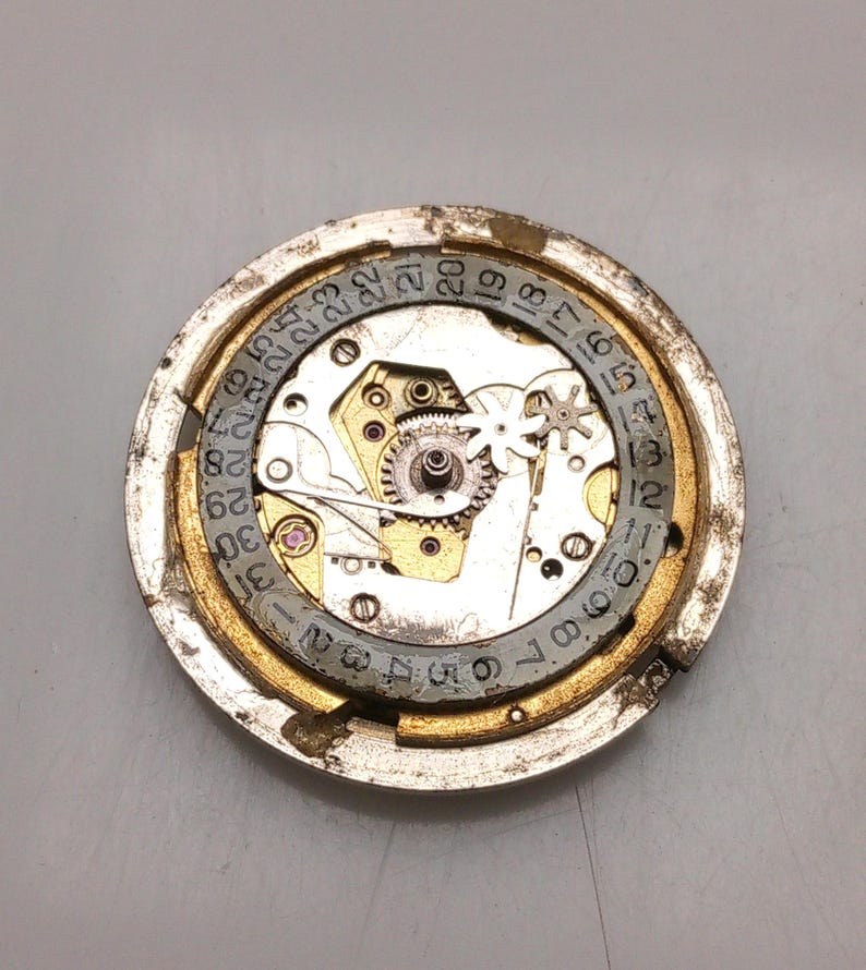 May include: A detailed view of a watch movement, showcasing intricate gears, springs, and a date dial. The circular mechanism features a gold-toned outer ring and a silver inner dial with numbered markings. The watch components are set against a grey background.
