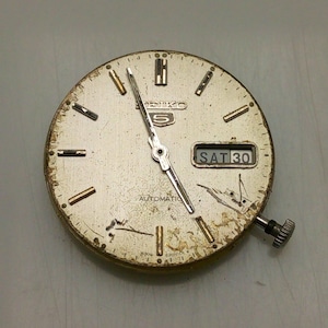 Może przedstawiać: Zegarek Seiko 5 w stylu vintage z srebrną tarczą, złotymi znacznikami godzin i wskaz&oacute;wkami. Dzień i data są wyświetlane w oknie na godzinie 3. Tarcza zegarka nosi ślady zużycia.