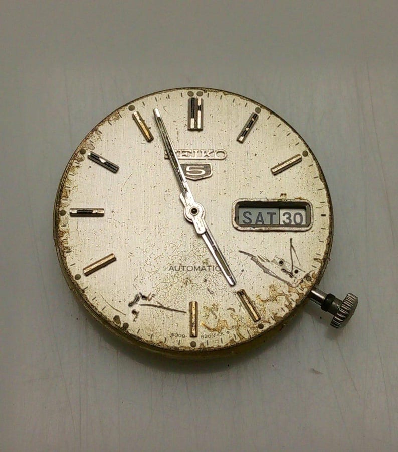 Może przedstawiać: Zegarek Seiko 5 w stylu vintage z srebrną tarczą, złotymi znacznikami godzin i wskaz&oacute;wkami. Dzień i data są wyświetlane w oknie na godzinie 3. Tarcza zegarka nosi ślady zużycia.