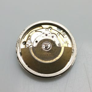 May include: Detailed view of a watch movement, showcasing the internal gears and mechanisms. The silver-toned metal casing houses the components, including a balance wheel and mainspring. The text "25 JEWELS SWISS" is visible.