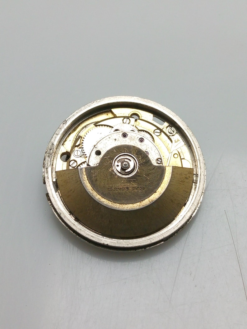 May include: Detailed view of a watch movement, showcasing the internal gears and mechanisms. The silver-toned metal casing houses the components, including a balance wheel and mainspring. The text "25 JEWELS SWISS" is visible.