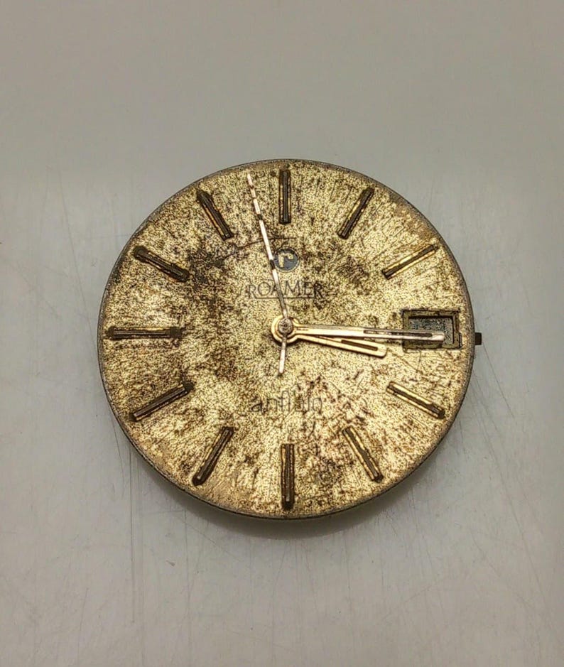 May include: A vintage gold-toned watch face with a textured surface. The watch face features gold-colored hour markers and hands. The brand name "ROAMER" is visible at the top. The watch face has a rectangular date window at the 3 o'clock position.