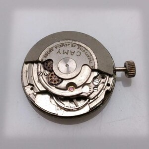May include: A close-up of a watch movement with the brand name "CAMY" visible. The movement is silver-toned with intricate gears and a small crown. The text "TWENTY-FIVE 25 JEWELS SWISS" is also visible.