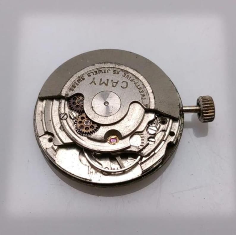 May include: A close-up of a watch movement with the brand name "CAMY" visible. The movement is silver-toned with intricate gears and a small crown. The text "TWENTY-FIVE 25 JEWELS SWISS" is also visible.