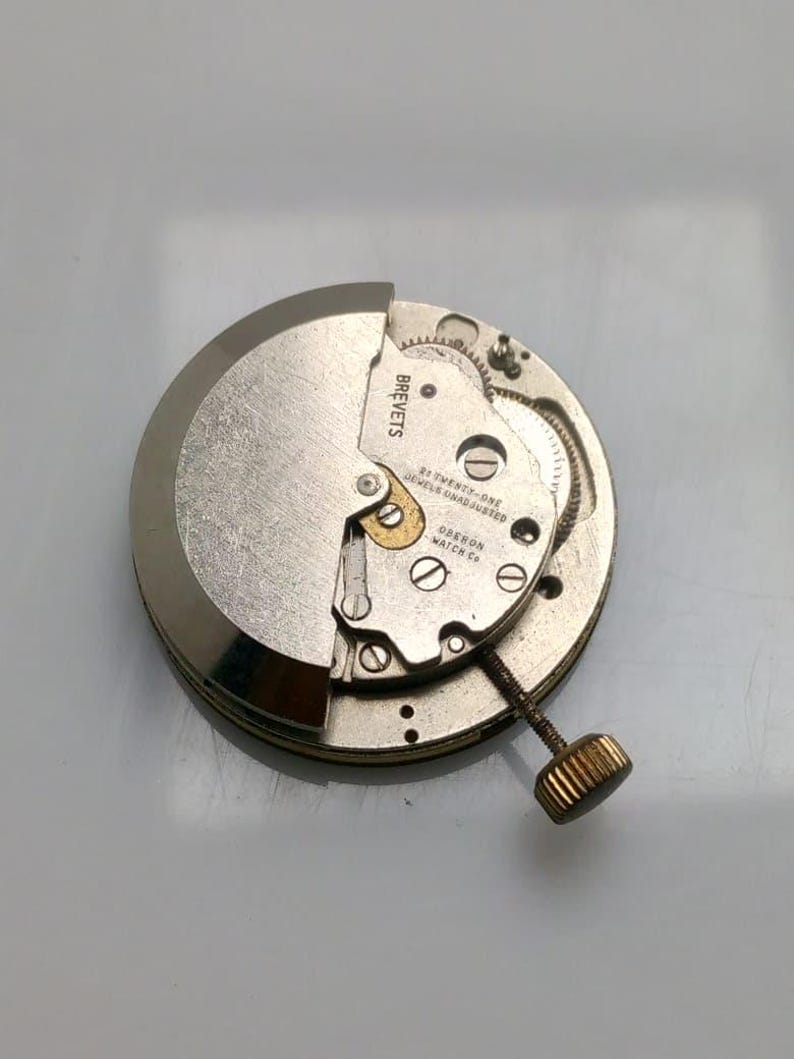 May include: A close-up of a watch movement with the text "BREVETS" and "OBERON WATCH CO." visible. The watch mechanism is silver and gold-toned, with intricate gears and a textured crown. The watch movement is a vintage item.