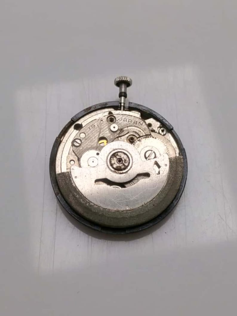 May include: A close-up of a watch movement with visible gears, screws, and a winding stem. The metal components are silver and gold, set within a black circular frame. The text "JAPAN" is visible on the movement.