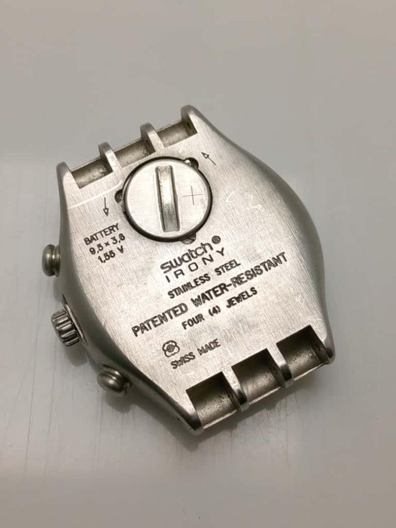 May include: A close-up of a Swatch Irony watch back. The stainless steel case features the text "PATENTED WATER-RESISTANT" and "FOUR (4) JEWELS". The watch back also includes the battery size: 9.5 x 3.6 mm, 1.55 V. Swiss made.
