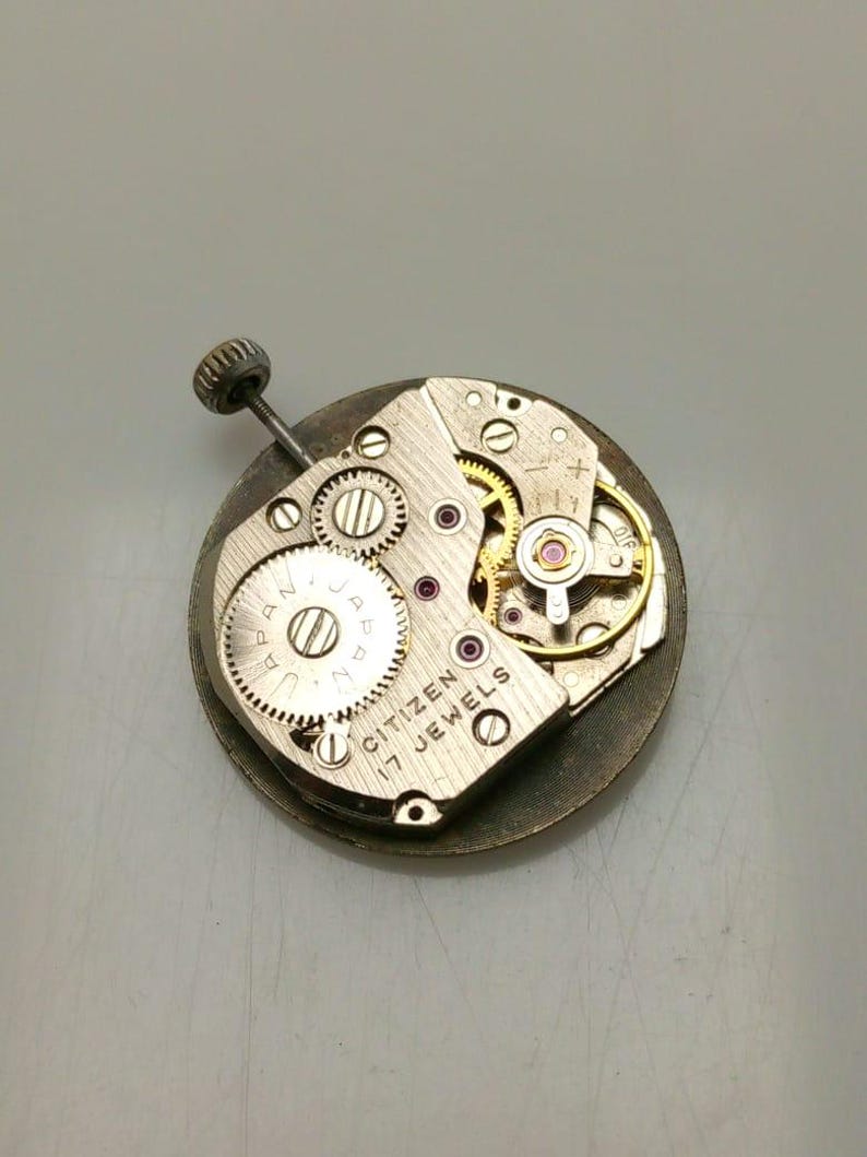 May include: Close-up of a Citizen 17 Jewels watch movement. The silver-toned mechanism features multiple gears, jewels, and a winding stem. The watch movement is circular and has the text "CITIZEN 17 JEWELS" engraved on it.