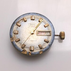 May include: A vintage Oberon automatic watch face with a silver dial, gold hands, and rectangular hour markers. The day and date are displayed in a window at the 3 o'clock position. The watch face has a gold crown and a blue and gold bezel.