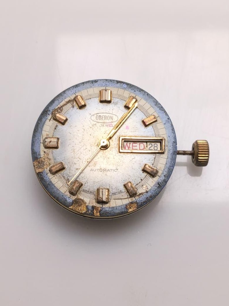 May include: A vintage Oberon automatic watch face with a silver dial, gold hands, and rectangular hour markers. The day and date are displayed in a window at the 3 o'clock position. The watch face has a gold crown and a blue and gold bezel.