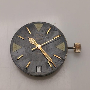 May include: A vintage watch face with a dark gray dial, gold-colored hands and markers, and a date window at the bottom. The watch face has the text "Ingersoll" and a textured appearance. A gold-colored crown is attached to the side.