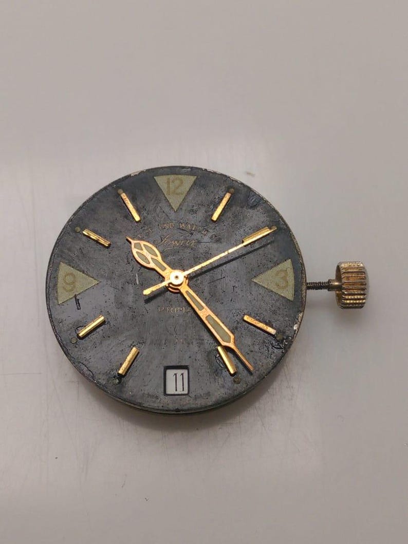May include: A vintage watch face with a dark gray dial, gold-colored hands and markers, and a date window at the bottom. The watch face has the text "Ingersoll" and a textured appearance. A gold-colored crown is attached to the side.