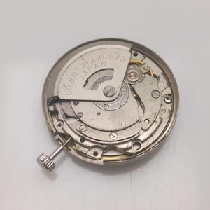 以下が含まれることがあります： 複雑な歯車とコンポーネントを備えた、銀色の時計ムーブメントのクローズアップ。金属プレートには「ORIENT 21J JAPAN 46943」の文字が見えます。小さなテクスチャード加工されたリューズが側面から出ています。