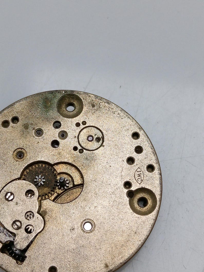 Pode incluir: Close-up de um movimento de rel&oacute;gio dourado com engrenagens vis&iacute;veis e a inscri&ccedil;&atilde;o "IWC" no lado.