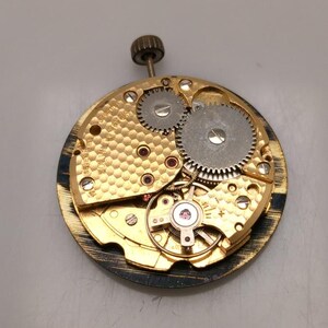 May include: Close-up of a vintage watch movement. The mechanism features gold-toned metal with intricate gears, ruby jewels, and a winding stem. The watch movement is circular and detailed.
