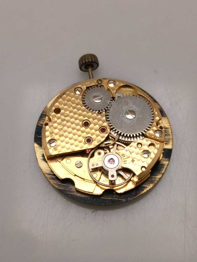 May include: Close-up of a vintage watch movement. The mechanism features gold-toned metal with intricate gears, ruby jewels, and a winding stem. The watch movement is circular and detailed.