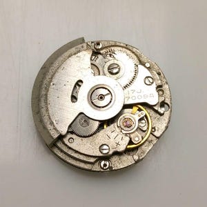 Może przedstawiać: Zbliżenie srebrnego mechanizmu zegarka z misternymi kołami zębatymi, śrubami i balansem. Widoczny jest tekst "17J 7009A". Okrągły mechanizm ma złożoną strukturę wewnętrzną.