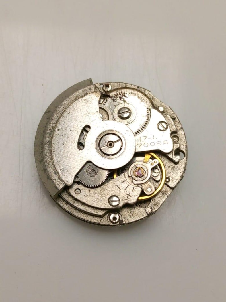 K&ouml;nnte beinhalten: Nahaufnahme eines silbernen Uhrwerks mit komplexen Zahnr&auml;dern, Schrauben und einer Unruh. Der Text "17J 7009A" ist sichtbar. Der kreisf&ouml;rmige Mechanismus zeigt eine komplexe Innenstruktur.