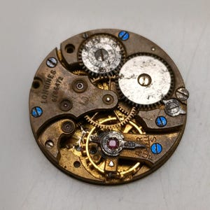 May include: Close-up of a vintage watch movement with intricate gears and components. The Longines brand name and serial number 6786472 are visible. The movement features a mix of gold, silver, and brass-colored parts, with blue accents.