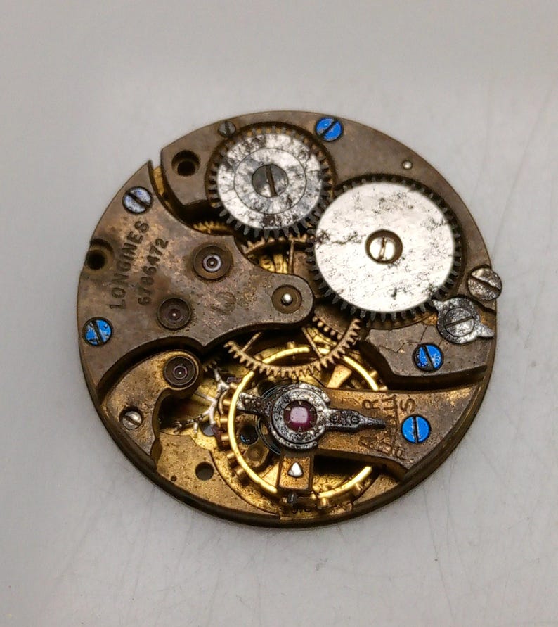 May include: Close-up of a vintage watch movement with intricate gears and components. The Longines brand name and serial number 6786472 are visible. The movement features a mix of gold, silver, and brass-colored parts, with blue accents.