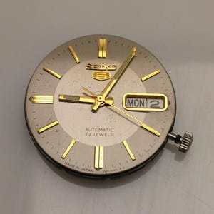 Pode incluir: Um mostrador de relógio Seiko 5 vintage com mostrador prateado e ponteiros e marcadores dourados. O dia e a data são exibidos em uma janela retangular. O mostrador do relógio diz "AUTOMATIC 23 JEWELS" e "MADE IN JAPAN".