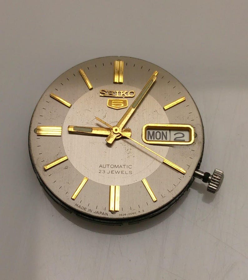 May include: A vintage Seiko 5 watch face with a silver dial and gold-tone hands and markers. The day and date are displayed in a rectangular window. The watch face reads "AUTOMATIC 23 JEWELS" and "MADE IN JAPAN".