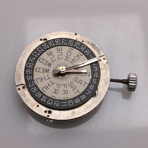 May include: A watch movement with a day and date display. The silver mechanism has a rotating dial with the days of the week and numbers 1-31. The hands are silver, and a small crown is visible. The measurements are in centimeters.