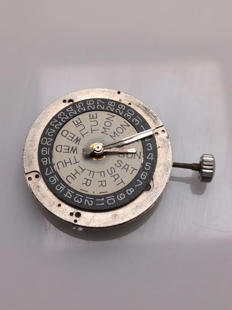 May include: A watch movement with a day and date display. The silver mechanism has a rotating dial with the days of the week and numbers 1-31. The hands are silver, and a small crown is visible. The measurements are in centimeters.