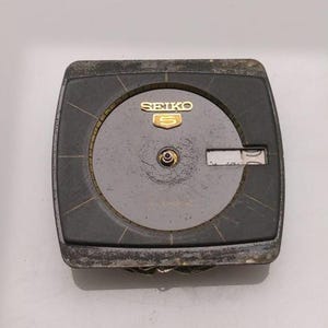 Puede incluir: Una esfera de reloj Seiko 5 vintage con un marco rectangular gris oscuro. La esfera del reloj es de color gris claro con agujas doradas y el logotipo de Seiko 5. La esfera del reloj muestra signos de desgaste.