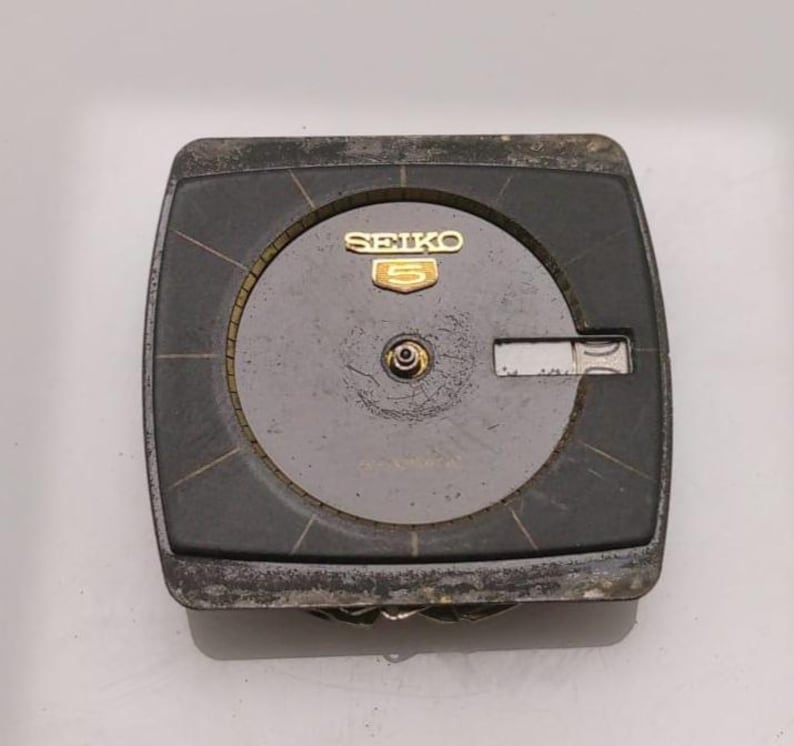 Puede incluir: Una esfera de reloj Seiko 5 vintage con un marco rectangular gris oscuro. La esfera del reloj es de color gris claro con agujas doradas y el logotipo de Seiko 5. La esfera del reloj muestra signos de desgaste.