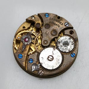 May include: Close-up of a vintage watch movement with intricate gears and components. The brass-colored mechanism features a ruby jewel and the text "LONGINES 6786472". Blue screws add contrast to the aged metal.