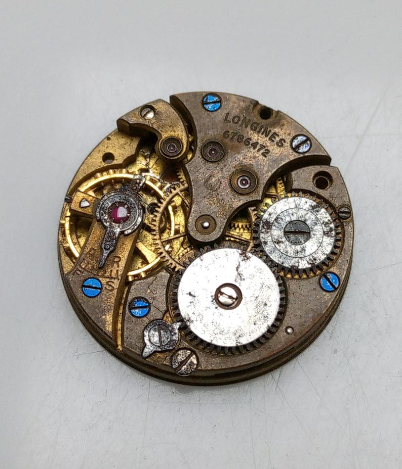 May include: Close-up of a vintage watch movement with intricate gears and components. The brass-colored mechanism features a ruby jewel and the text "LONGINES 6786472". Blue screws add contrast to the aged metal.