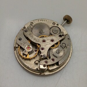 Op de afbeelding: Een zilveren uurwerk met een gouden opwindstift en de tekst "18 Jewels" en "35009".
